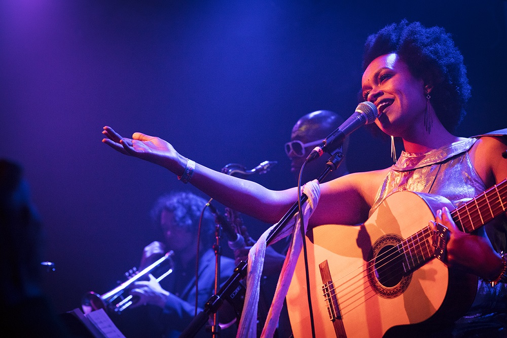 Meklit Hadero on stage with guitar and in front of a mic stand