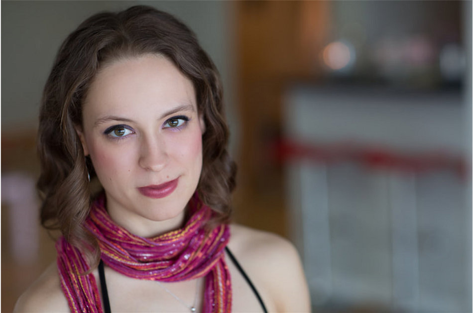 Lisa Neher is situated on the left hand side with a blurry background. She has short, curly brown hair, simple yet noticeable makeup, and a pink scarf.